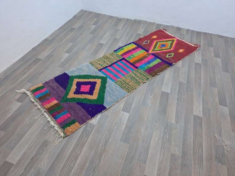 May include: A colorful, geometric patterned rug with a long, rectangular shape. The rug features a variety of colors, including red, pink, blue, green, and yellow. The rug is made of a soft, woven material and has a fringed edge.