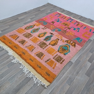 May include: A colorful rug with a geometric pattern in shades of pink, orange, brown, and blue. The rug has a fringe border and is made of a soft, fluffy material.