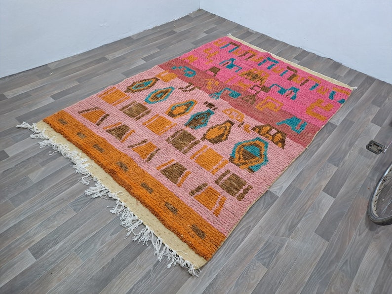 May include: A colorful rug with a geometric pattern in shades of pink, orange, brown, and blue. The rug has a fringe border and is made of a soft, fluffy material.