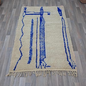 May include: A white wool rug with a blue geometric design. The design features two tall, thin figures with a zig-zag pattern at the bottom.