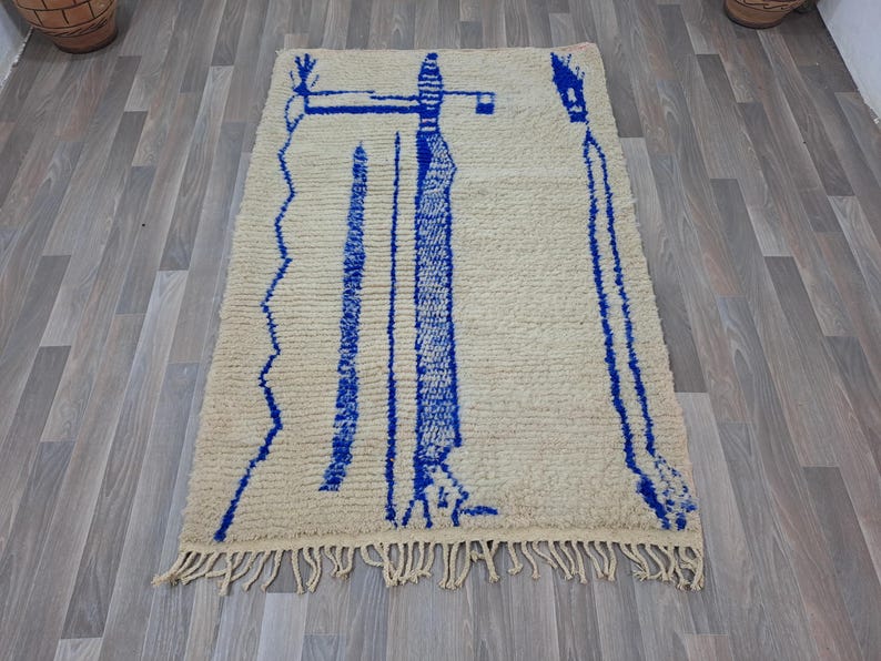 May include: A white wool rug with a blue geometric design. The design features two tall, thin figures with a zig-zag pattern at the bottom.