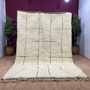 Puede incluir: Una alfombra de lana blanca con un patrón geométrico negro. La alfombra tiene un borde de flecos y está colocada sobre un suelo de madera.