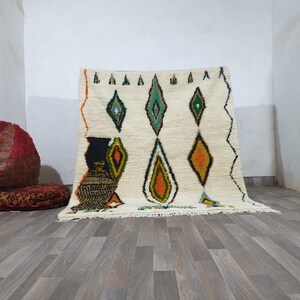 May include: Cream-colored, hand-woven rug with a diamond pattern in green, orange, and black. The rug has a fringed edge and is displayed against a white wall and wooden floor. The design is geometric and the rug is likely made of wool.