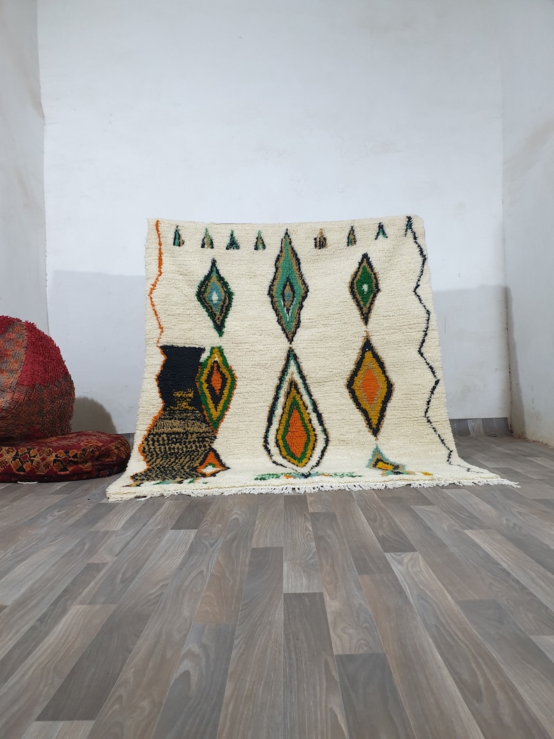 May include: Cream-colored, hand-woven rug with a diamond pattern in green, orange, and black. The rug has a fringed edge and is displayed against a white wall and wooden floor. The design is geometric and the rug is likely made of wool.