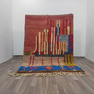 May include: A colourful, hand-woven rug with an abstract design. The rug has a red background with vertical stripes in orange, white, and blue. A blue and yellow border with fringe adds a decorative touch. The rug is made of wool.