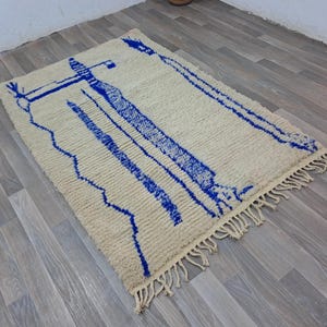 May include: A white and blue Moroccan rug with a geometric pattern. The rug has a fringe border and is made of wool.