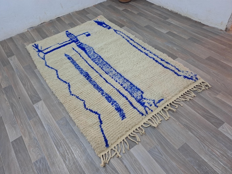 May include: A white and blue Moroccan rug with a geometric pattern. The rug has a fringe border and is made of wool.