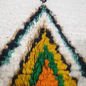 May include: Close-up of a white rug with a geometric pattern. The design features a central diamond shape in yellow, orange, and green, outlined in black. Additional teal and tan accents are visible.