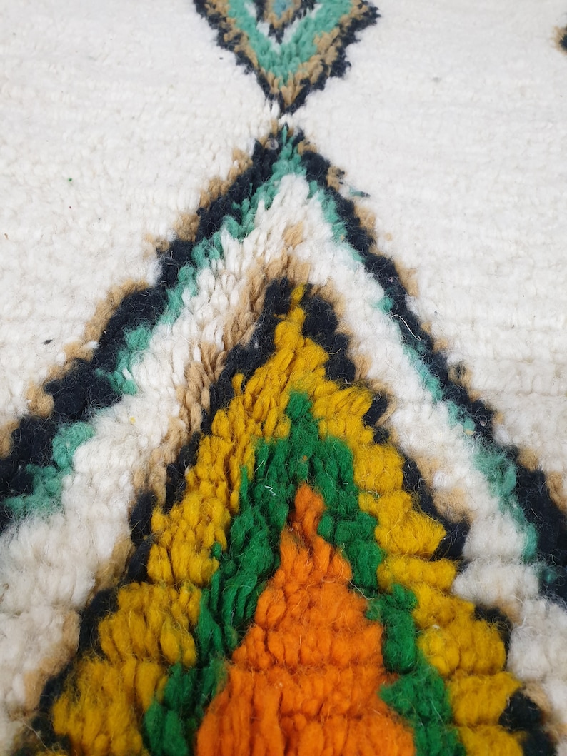 May include: Close-up of a white rug with a geometric pattern. The design features a central diamond shape in yellow, orange, and green, outlined in black. Additional teal and tan accents are visible.