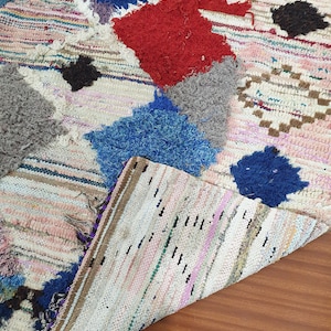 May include: A white, handwoven rug with colorful patches of blue, red, gray, and black. The rug has a striped pattern and is made of recycled materials.