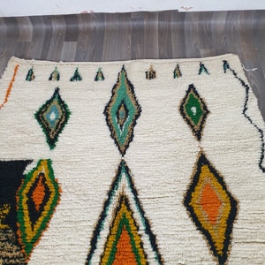 May include: Cream-colored rug with a geometric diamond pattern in shades of green, orange, yellow, and black. The rug has a textured, woven appearance, with a white background and a border of small green triangles.