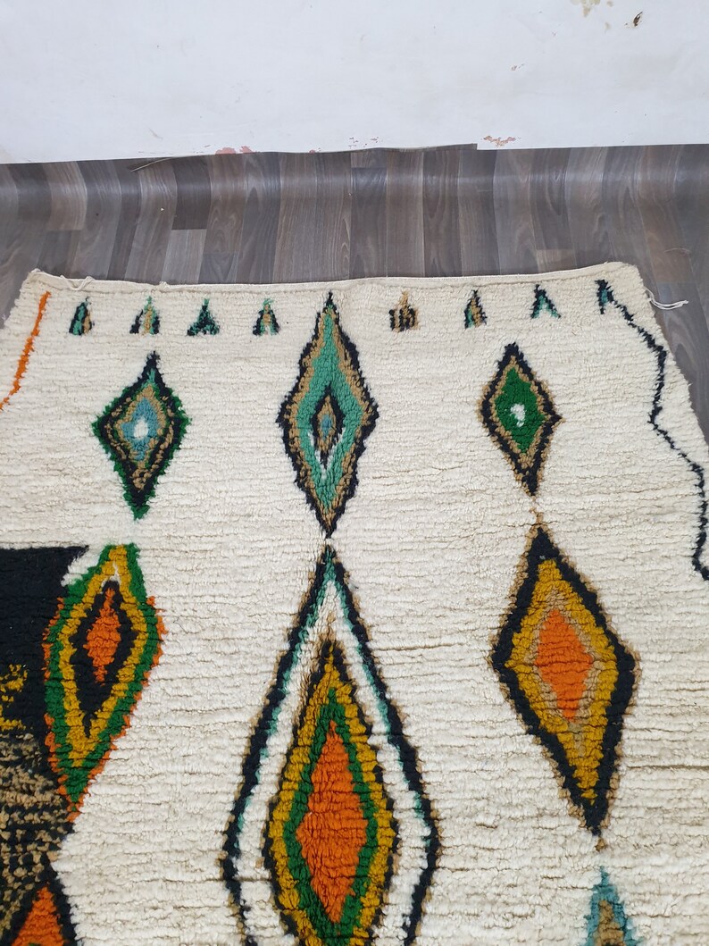 May include: Cream-colored rug with a geometric diamond pattern in shades of green, orange, yellow, and black. The rug has a textured, woven appearance, with a white background and a border of small green triangles.