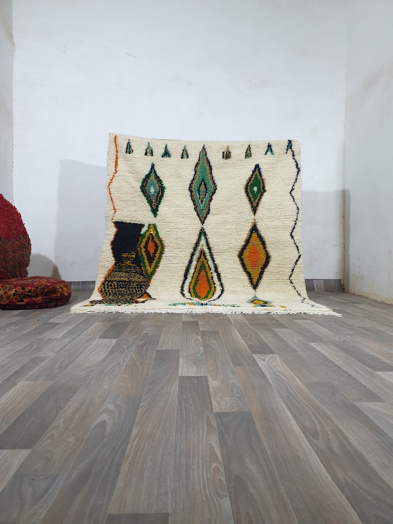 May include: Cream-colored, hand-woven rug with a diamond pattern in green, orange, and black. The rug has a fringed edge and a geometric design. The rug is placed on a wooden floor.