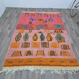 May include: A colorful Moroccan rug with a geometric pattern in shades of pink, orange, brown, and blue. The rug has a fringe border and is made of wool.