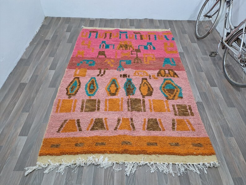 May include: A colorful Moroccan rug with a geometric pattern in shades of pink, orange, brown, and blue. The rug has a fringe border and is made of wool.