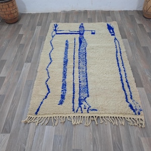 May include: A white and blue Moroccan rug with a geometric design. The rug has a fringe border and is made of wool.
