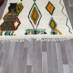 May include: A white rug with a geometric diamond pattern in shades of green, orange, and black. The rug has a fringe along the bottom edge and is placed on a wooden floor.
