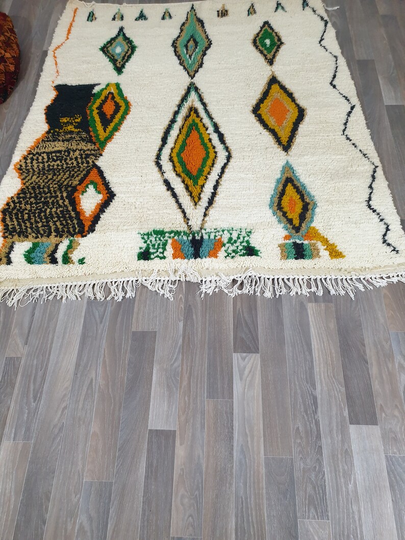May include: A white rug with a geometric diamond pattern in shades of green, orange, and black. The rug has a fringe along the bottom edge and is placed on a wooden floor.
