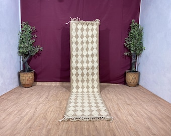 May include: A white and beige runner rug with a diamond pattern. The rug has a fringe border and is laying on a wooden floor.