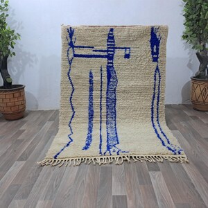 May include: A white and blue Moroccan rug with a geometric pattern. The rug has a fringe border and is made of wool.