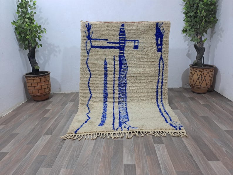 May include: A white and blue Moroccan rug with a geometric pattern. The rug has a fringe border and is made of wool.