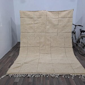 May include: A large, off-white, rectangular rug with a geometric pattern of squares. The rug has a fringe border and is lying on a wooden floor.