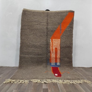 Peut inclure: Un tapis rectangulaire à la surface en laine brune texturée, orné d'un grand motif graphique abstrait aux tons orange vif, blanc et bleu. Le tapis est orienté verticalement et présente des franges de couleur crème sur le bord inférieur.