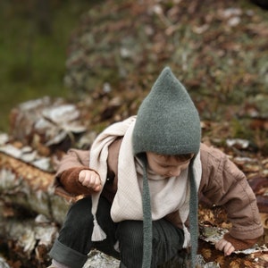 以下が含まれることがあります： 耳当て付きのグレーのニット帽、クリーム色のスカーフ、茶色のジャケット、コーデュロイパンツを着た子供。 子供は丸太の上に座っており、ベージュのブーツを履いています。