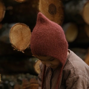 Może przedstawiać: Dziecko w brązowej pikowanej kurtce i bordowym dzianinowym berecie z ostrym czubkiem. Dziecko stoi przed stosami ułożonych kłód.