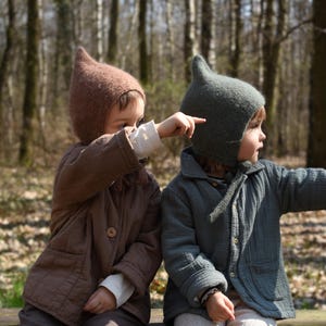 Könnte beinhalten: Zwei Kinder tragen passende Strickmützen mit spitzen Spitzen und Jacken mit Knöpfen. Eine Mütze ist braun, die andere graugrün. Die Kinder sitzen auf einer Holzbank in einem Wald und zeigen in die gleiche Richtung.