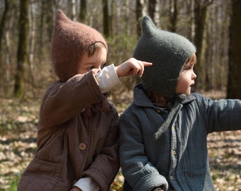May include: Two children wearing matching knit hats with pointed tops and button-up jackets. One hat is brown, the other is gray-green. The children are sitting on a wooden bench in a forest, pointing in the same direction.