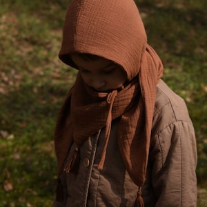 May include: A child wearing a brown hooded scarf and a brown jacket. The scarf has tassels on the ends.