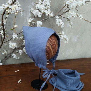 May include: A blue bonnet and bib set made from a soft, textured fabric. The bonnet has ties under the chin, and the bib is square with a rounded neck and ties. The set is displayed on a wooden surface, with a floral background.