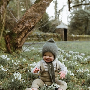 Op de afbeelding: Een lachende baby draagt een groene gebreide muts, bijpassende sjaal en kaki tuinbroek. De outfit van de baby omvat een shirt met lange mouwen en bruine en witte schoenen. De achtergrond toont bomen en een kleine stenen structuur.