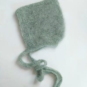 May include: A sage green knitted baby bonnet with long ties. The bonnet is made of a fuzzy, lightweight material and is designed to cover the head and tie under the chin. The bonnet is a light green colour.