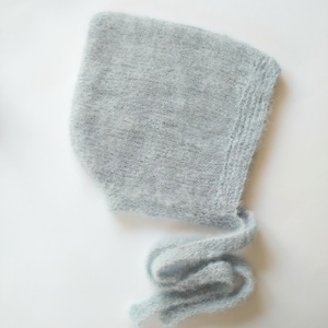 Peut inclure: Un bonnet tricoté bleu clair avec de longues attaches. Le bonnet est fait d'une matière douce et duveteuse et a une forme arrondie qui couvre la tête et les oreilles. Les attaches sont longues et peuvent être nouées sous le menton. Le bonnet est probablement pour un bébé ou un jeune enfant.