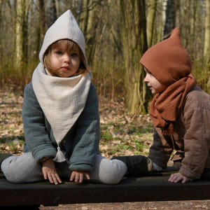 Op de afbeelding: Twee kinderen dragen gekapte sjaals, een in beige en een in bruin, zitten op een houten bank in een bos.