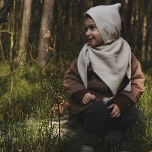 Peut inclure: Un jeune enfant portant une veste marron et une écharpe à capuche blanche est assis sur une bûche dans une forêt. L'enfant regarde vers la droite du cadre.