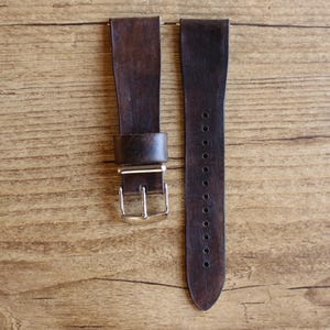 Puede incluir: Una correa de reloj de cuero marrón oscuro de dos piezas con un acabado liso y mate. La correa incluye una hebilla de metal plateado y una trabilla de cuero a juego, con una serie de orificios de ajuste circulares en la pieza larga.