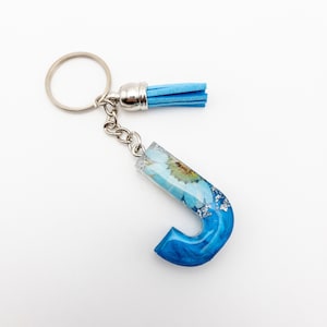 May include: A blue resin letter J keychain with a dried flower embedded in the resin. The keychain has a silver ring and a blue tassel.