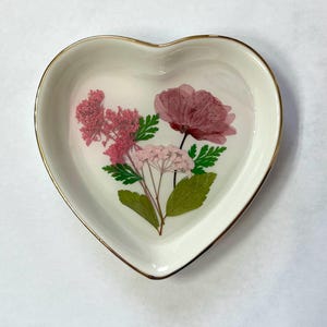 Könnte beinhalten: Eine weiße herzförmige Schale mit goldenem Rand, verziert mit gepressten rosa Blüten und grünen Blättern.