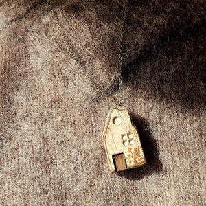 Pode incluir: Colar com um pingente de casa de madeira. O pingente é uma casa em miniatura com uma pequena porta, janelas e um design decorativo de árvore. A corrente do colar é de cor ouro antigo.