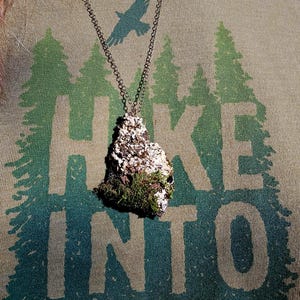 May include: A pendant necklace with a natural, organic design. The pendant is a rough, irregular shape, covered in moss and small white accents. The necklace chain is a dark, antique metal. The pendant hangs against a green t-shirt with a forest graphic and the words "Hike Into The".