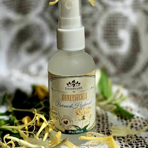 Puede incluir: Una botella de spray de vidrio transparente de Honeysuckle Botanical Hydrosol con una tapa blanca, adornada con flores amarillas de madreselva. La etiqueta dice "Vonderwoods Honeysuckle Botanical Hydrosol" y "Handmade with Love". La botella contiene 59 ml / 2.0 fl oz.
