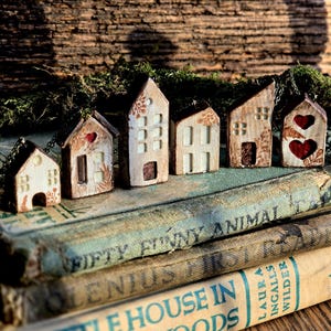 May include: A row of small, decorative wooden houses with unique features such as heart cutouts and floral motifs. They are displayed on old books, including one titled "Little House in the Big Woods". The backdrop is a textured wooden surface, creating a rustic aesthetic.