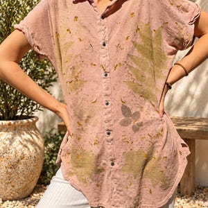 May include: A dusty pink button-down shirt featuring short sleeves, a classic collar, and a botanical print with olive green leaf patterns and speckled details. The garment has a curved hemline and dark buttons running down the center front.