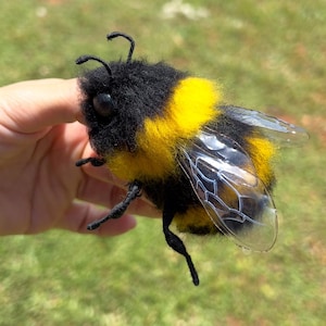 Puede incluir: Una figura de abejorro hecha a mano con un cuerpo negro y amarillo, alas transparentes y pequeñas patas y antenas negras. La abeja se sostiene en una mano sobre un fondo verde borroso.