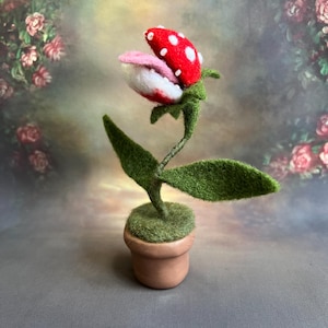 May include: A felt sculpture of a carnivorous plant with a red and white mouth, green leaves, and a brown pot. The plant is a popular video game character.