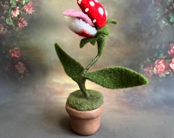 May include: A felt sculpture of a carnivorous plant with a red and white mouth, green leaves, and a brown pot. The plant is a popular video game character.