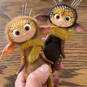 Puede incluir: Dos muñecas de fieltro hechas a mano con cuerpos marrones y dorados, ojos grandes y bufandas de punto amarillo. Las muñecas tienen manchas blancas y se abrazan.